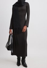 Robe longue noire à côtes avec manches longues, associée à une écharpe noire texturée, tenant un sac à main noir et portant des bottines noires.