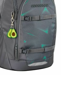 Sac à dos gris avec des motifs géométriques verts, comportant un porte-clés en forme de deux moitiés d'avocat attaché à une fermeture éclair et une sangle latérale.