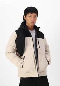 Beige und schwarze Steppjacke mit einem Reißverschluss an der Vorderseite, großen Taschen und einer abnehmbaren Schultertasche. Kapuzen-Design mit elastischen Bündchen.