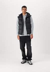 Chaleco puff negro superpuesto a una sudadera gris, combinado con jeans oscuros y zapatillas plateadas. Textura suave y diseño acolchado.