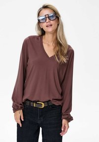 Bruine langemouwen V-hals top met elastische mouwdetails, gecombineerd met zwarte high-waisted jeans en een donkere riem met een gouden gesp.