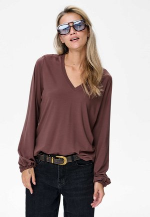 Braunes langärmeliges V-Ausschnitt-Oberteil mit elastischen Bündchen, kombiniert mit schwarzen hochtaillierten Jeans und einem dunklen Gürtel mit einer goldenen Schnalle.