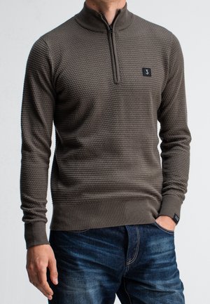 Homme portant un pull gris foncé texturé à fermeture éclair quart, et un jean bleu, avec une main dans la poche sur un fond uni.