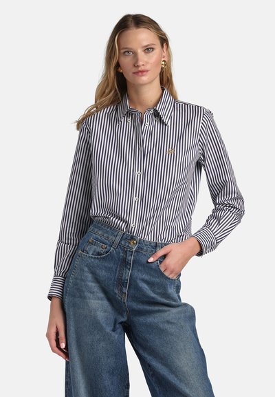 Plava žena s zlatnim naušnicama, plavo-bijelom košuljom s vertikalnim prugama i visokim strukom širokim plavim trapericama, s jednom rukom u džepu.