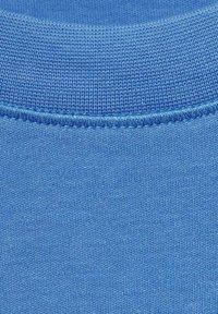 Tastă de bumbac albastră tricotată, cu un decolteu rotund ribat. Textură netedă cu cusături uniform dispuse și o ușoară strălucire.