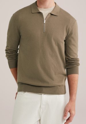 Homme portant un pull polo zippé à manches longues vert olive et un pantalon blanc, debout avec une main derrière le dos et l'autre détendue.
