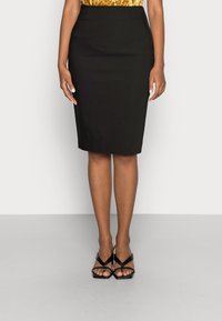 Femme portant une jupe crayon noire jusqu'aux genoux, un top doré et des sandales à talons hauts noires à lanières, se tenant contre un fond blanc.