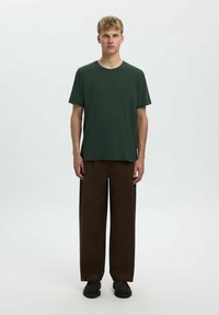 Camiseta de algodón verde oscuro con un corte relajado, combinada con pantalones marrones holgados. El modelo está de pie frente a un fondo neutro.