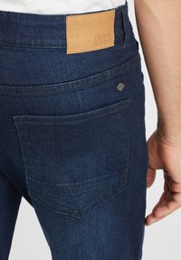 Jeans de mezclilla azul oscuro con un parche de cuero etiquetado "EL VIAJE DEL DENIM", que presentan un bolsillo trasero y una tela texturizada.
