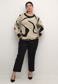 Pull beige avec des motifs abstraits noirs, col rond, manches longues. Associé à un pantalon sombre et des escarpins noirs à bouts pointus. Texture douce.