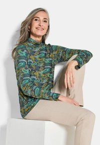 Langärmliges Oberteil mit Paisley-Muster in Grün- und Blautönen, ausgestattet mit einem enganliegenden Stehkragen und glattem Material, kombiniert mit hellbeigen Hosen.