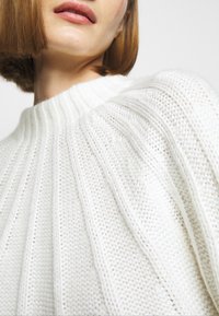 Femme aux cheveux courts châtain clair portant un pull en maille côtelée blanche, montrant la zone du cou et des épaules sur un fond clair uni.