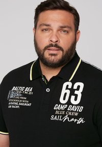 Black polo shirt with a contrasting yellow collar, featuring white embroidered text and numbers, textured fabric, and button closure.