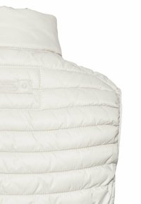 Leichte, cremefarbene Pufferweste mit horizontalen Nähten, hohem Kragen und einem kleinen Etikett auf der Rückseite. Glatte, gesteppten Textur.