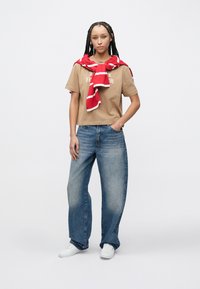 Μπεζ βαμβακερό t-shirt με το κείμενο "TOMMY HILFIGER", συνδυασμένο με ανοιχτό μπλε παντελόνι φαρδιάς γραμμής και κόκκινο και λευκό ριγέ κασκόλ που είναι περασμένο γύρω από το λαιμό.