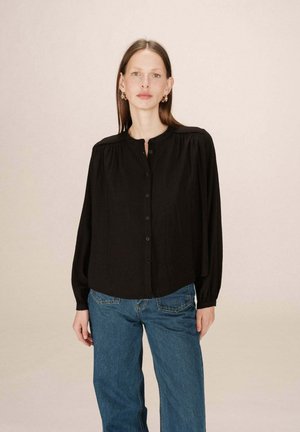 Chemise noire à manches longues, boutonnée, avec un col arrondi et des détails froncés aux épaules. Portée avec un jean bleu taille haute. Texture de tissu lisse.