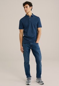 Tamsiai mėlynas polo marškinėlis su tekstūrine raštika, dviejų mygtukų užsegimu, derinamas su mėlynomis džinsinėmis kelnėmis ir pilkšvomis sportbačiais, stovintis neutraliame fone.