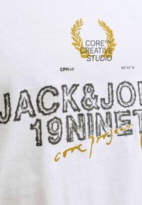 Camiseta blanca con texto y gráficos impresos en negro y dorado, incluyendo "JACK&JON" y "19NINET", con un diseño de corona de laurel.