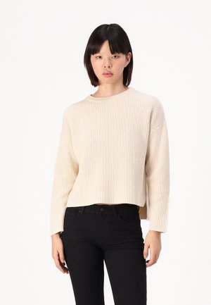 Beige ribbestrikket genser med rund hals og senkede skuldre, kombinert med svarte tettsittende jeans; teksturen er myk og tykk.