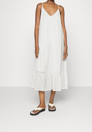 Femme portant une robe midi blanche fluide sans manches avec des bretelles fines et un ourlet volanté, assortie à des sandales à plateforme blanches avec semelles noires.
