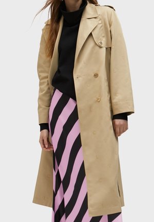 Vrouw draagt een beige trenchcoat over een zwarte coltrui en een roze-zwart diagonale gestreepte rok, staand tegen een neutrale achtergrond.