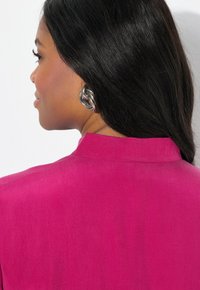 Fuchsia blouse met kraag, gladde textuur, en een achteraanzicht van grote metalen oorbellen, met lang donker haar dat over de schouder valt.
