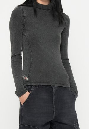 Personne portant un haut moulant côtelé à manches longues gris foncé et un pantalon ample gris foncé avec les mains dans les poches, debout sur un fond blanc.