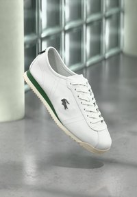 Λευκό δερμάτινο αθλητικό παπούτσι με πράσινη λωρίδα στη σόλα, κορδόνια και μαύρο λογότυπο κροκοδείλου, τοποθετημένο μπροστά σε φόντο από ανακλαστικό τοίχο από γυάλινα τούβλα.