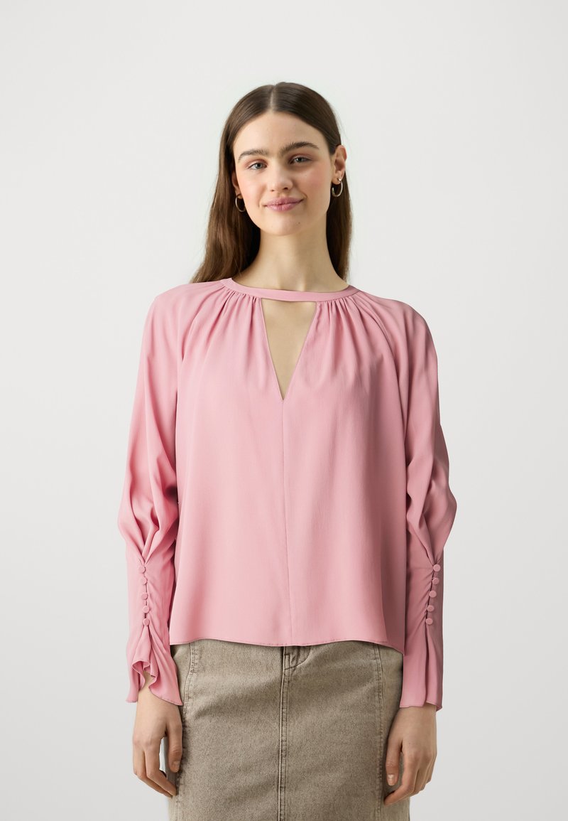 Blusa rosa chiaro con scollatura a fessura, dettagli arricciati e maniche lunghe con bottoni ai polsini. Tessuto morbido.