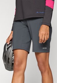 Graue Shorts aus leichtem Material, mit flachen Nähten, Seitentaschen und einem Logo am unteren Bein. Ein Helm wird in einer Hand gehalten.