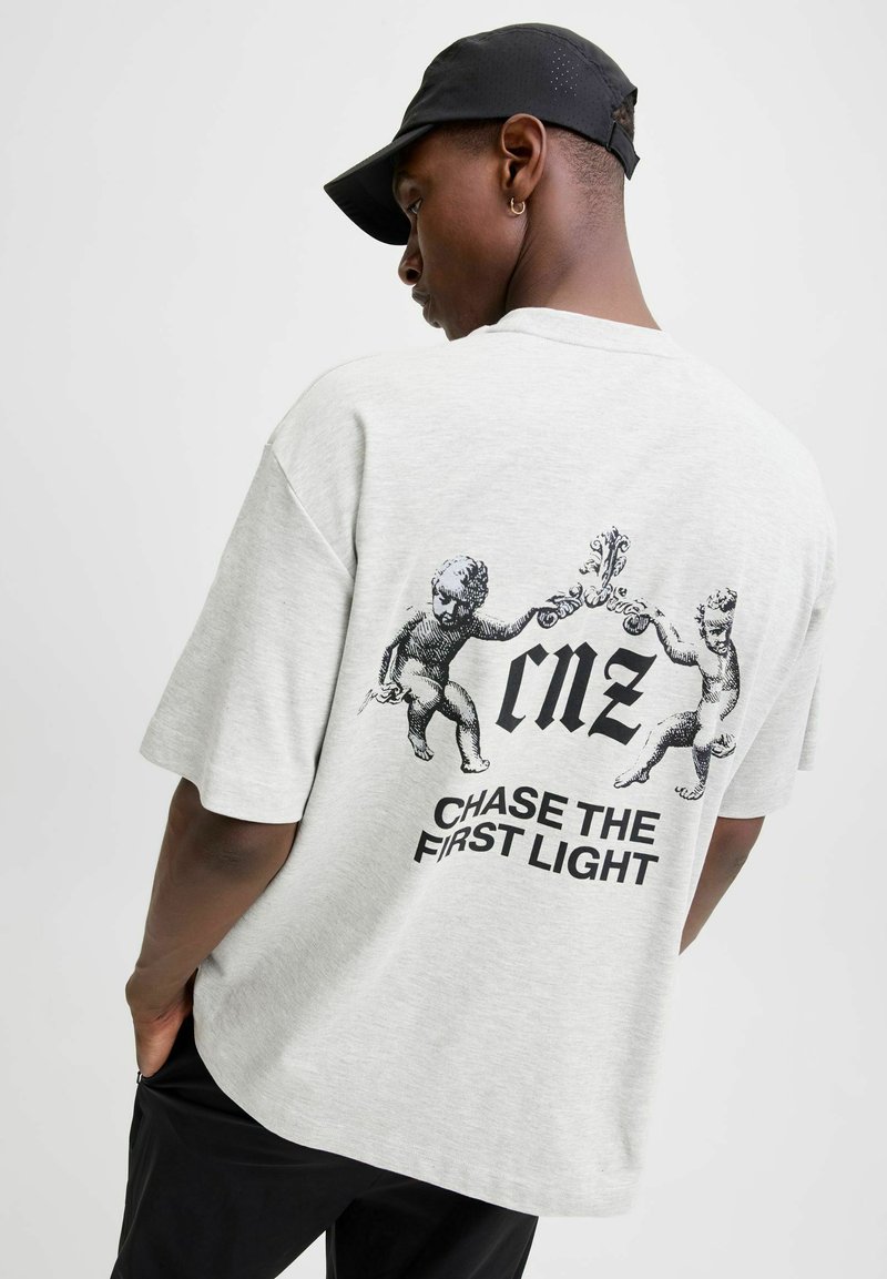 Lys grå bomulds t-shirt med en afslappet pasform. Har et grafisk print af engle og teksten "cmz CHASE THE FIRST LIGHT" på ryggen.