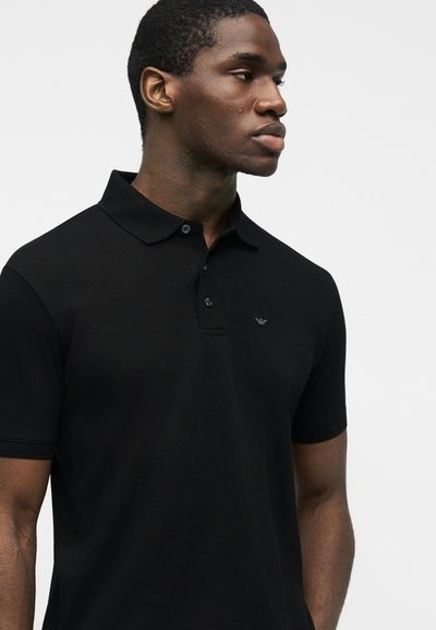 Νεαρός μαύρος άνδρας που φοράει μαύρο πόλο με κοντά μανίκια, κουμπιά και ένα μικρό λογότυπο, κοιτάζοντας προς τα δεξιά μπροστά σε απλό φωτεινό φόντο.