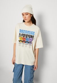 Krämfärgad oversized t-shirt med en grafisk tryck av färgglada tecknade figurer. Kompletterad med blå denim cargo-byxor och en vit mössa.