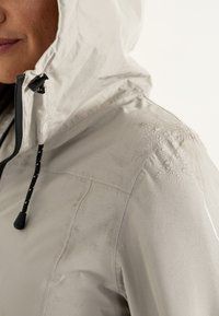 Wasserabweisende Kapuzenjacke in Hellgrau mit strukturiertem Finish, schwarzem Kordelzug und dezenten Wassertropfen auf der Schulter.