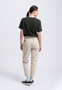 Donkergroene katoenen T-shirt, beige broek met vijf zakken en stikdetails, zwarte sneakers en pastelgekleurde sokken. Model geposeerd van achteren.