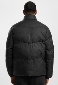 Sort polstret jakke med høj krave, med et quiltet design, detaljer ved fronten og elastiske manchetter. Glat, blank stoftekstur.