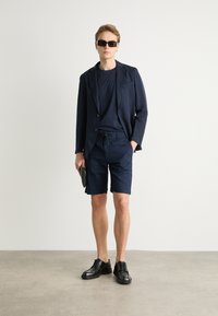 Blazer blu navy su una camicia blu navy, abbinato a pantaloni corti blu navy. Scarpe in pelle nere. Il modello tiene una pochette nera, indossando occhiali da sole scuri.