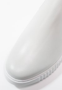 Chaussure en cuir blanc à enfiler avec une finition lisse, bout arrondi, et semelle en caoutchouc texturée avec des nervures verticales.