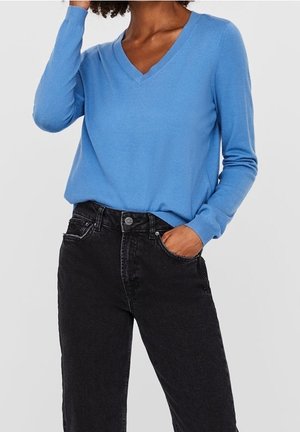 Personne portant un pull bleu clair à col en V et un jean noir taille haute, une main dans la poche et l'autre touchant ses cheveux.