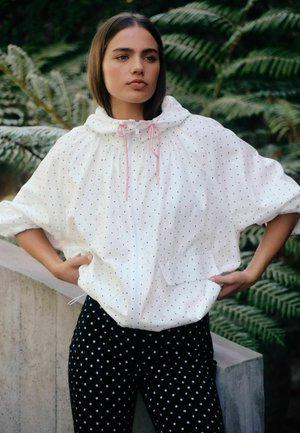 Junge Frau steht im Freien und trägt eine weiße Jacke mit rosa Kordeln und schwarzen Punkten sowie eine schwarze Hose mit weißen Punkten, vor einem Hintergrund aus grünen Farnen.