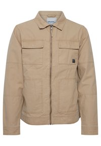 Beige, glidelåsjakke laget av bomull med krage, to brystlommer og et logo-patch på venstre side. Har en avslappet passform.