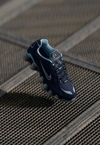 Tumši zili Nike Shox sporta apavi ar sudraba akcentiem un mīksto zoli uz teksturētas metāla režģa virsmas.