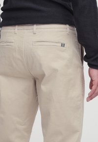 Beige pants with two back welt pockets, one featuring a small black and white striped fabric detail, worn with a black textured sweater.