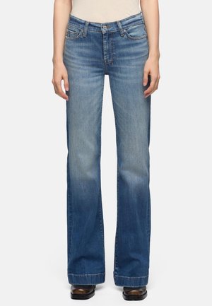 Kvinde iført blåtonede, brede jeans med falmet look og brune sko med firkantet tå, stående mod en ensfarvet hvid baggrund.