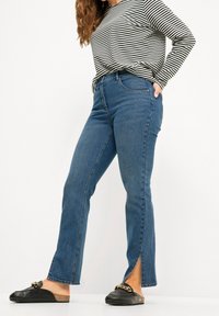 Jeans de mezclilla oscuros de tiro alto con corte recto y pierna ligeramente acampanada, que cuentan con aberturas laterales en el dobladillo. Llevados con una blusa de rayas y mocasines negros.