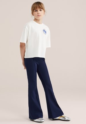 Mladić u bijeloj majici kratkih rukava s plavim logotipom i tamnoplavim hlačama proširenog kroja, stoji ispred jednobojne svijetle pozadine.