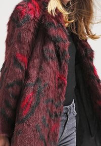 Abrigo de piel sintética color carmesí con un patrón llamativo en negro y rojo, mangas largas y textura suave, combinado con una camiseta negra y vaqueros grises.