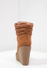 Brązowy zamszowy botek za kostkę z zaokrąglonym wierzchołkiem, pikowanym brązowym akcentem wokół kołnierza oraz teksturowanym koturnem.