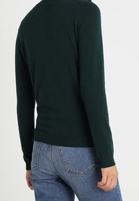 Person wearing a dark green long-sleeve sweater and blue jeans, shown from behind against a plain white background.