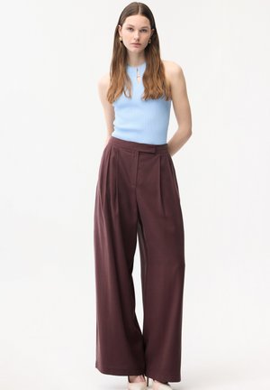 Kvinde iført en ærmeløs lyseblå ribstrikket top og højtaljede, vidtbenede mørkebrune bukser, stående med hænderne bag ryggen.